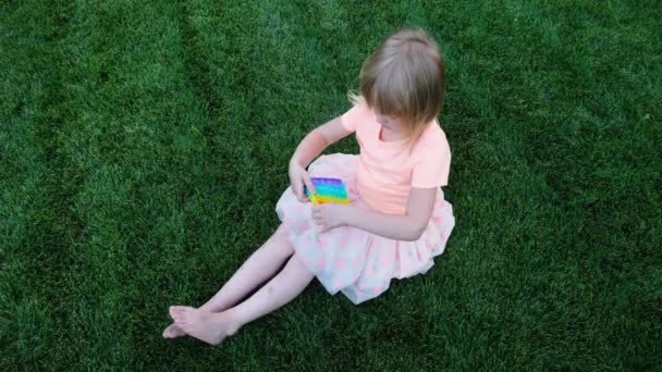 Blond klein meisje zitten op het gras spelen met pop het duwen pop bubble fidget speelgoed. — Stockvideo