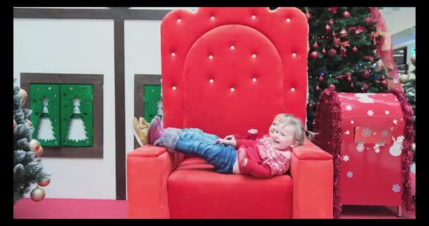 Dwie radosne dziewczynki pobłażają wielkiemu czerwonemu krzesłu Świętego Mikołaja i czekają, aż je odwiedzi. — Wideo stockowe