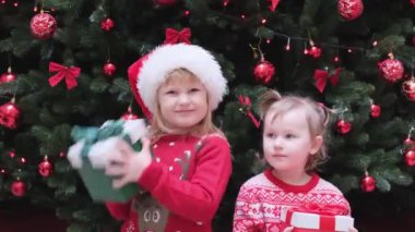 İki küçük kız Noel ağacının yanında durur ve içinde ne olduğunu öğrenmek için hediye kutusunu sallar..
