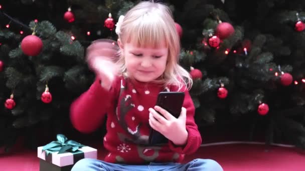 Menina loira sorridente usando telefone inteligente conversando com a família parabeniza Feliz Natal por videochamada. — Vídeo de Stock
