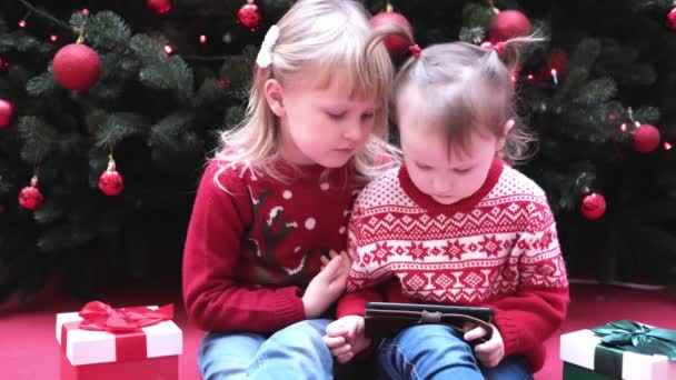 Kleine Mädchen Schwestern sitzen in der Nähe des Weihnachtsbaums und beobachten den Bildschirm, lesen, schreiben, spielen. — Stockvideo