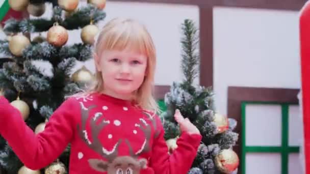 크리스마스 스웨터를 입은 작은 소녀가 손으로 손바닥을 흔들며 인사의 몸짓으로 누군가를반긴다 — 비디오