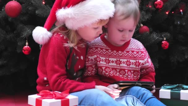 Kleine meisjes zussen zitten in de buurt van de kerstboom en kijken naar scherm, lezen, typen, het spelen van spelletjes. — Stockvideo