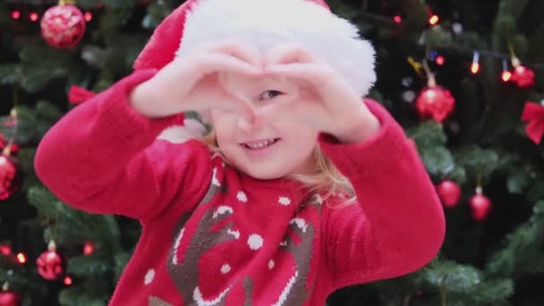 Happy Little Girl stanąć w pobliżu choinki w czerwonym kapeluszu Santa zrobić serce z rąk — Wideo stockowe