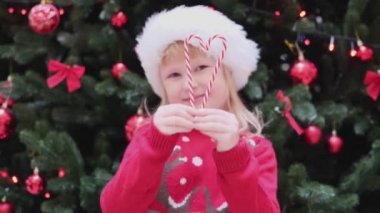 Kırmızı Noel Baba şapkalı Mutlu Küçük Kız Noel ağacının yanında durur şeker kamışından kalp yapar