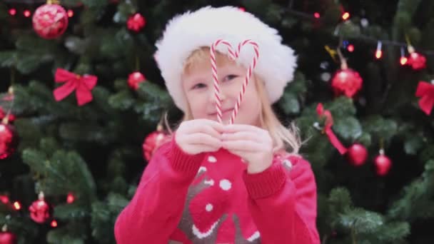 Happy Little Girl de pie cerca del árbol de Navidad en el sombrero de santa roja hacer corazón de bastón de caramelo — Vídeo de stock