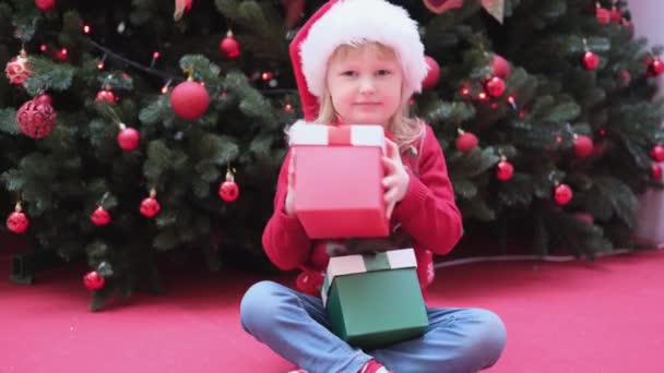 Mała dziewczynka siedzi w pobliżu choinki i potrząsa pudełko z prezentami, aby dowiedzieć się, co jest w środku. — Wideo stockowe