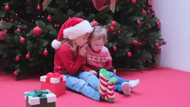 Sorelle bambine sedute vicino all'albero di Natale e guardando per schermare, leggere, digitare, giocare. — Video Stock