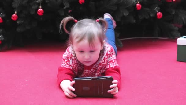 크리스마스 트리 근처의 작은 소녀가 화면을 보고, 책을 읽고, 타자치고, 게임을 하고 있습니다.. — 비디오