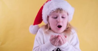 Noel Baba şapkalı güzel sarışın kız kameraya altın parıltılı konfeti üflüyor ve gülümsüyor, sarı stüdyo geçmişi var.
