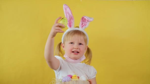 Porträtt av glad blond liten flicka leker med påskägg på gul bakgrund. — Stockvideo