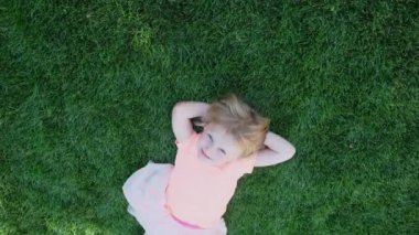 Happy little blond girl lies on green grass. The child is dream on the grass in park.