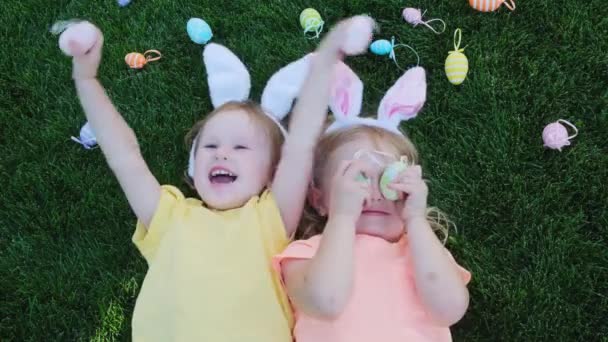 Щасливі маленькі дівчата, сестри розважаються на траві, закривають очі кольоровими великодніми яйцями . — стокове відео