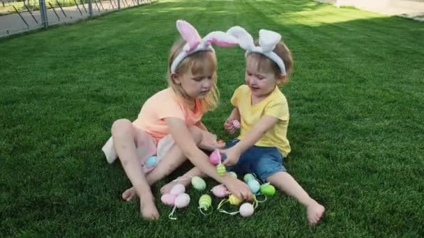 Hermanitas felices sentadas en la hierba verde y jugando con huevos de Pascua . — Vídeo de stock
