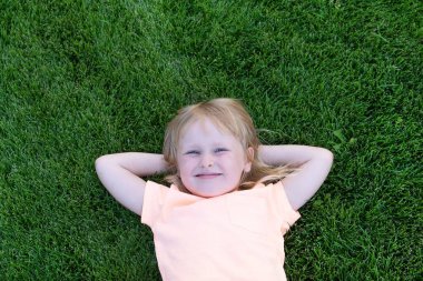 Yeşil çimlerin üzerinde tişörtlü mutlu küçük sarışın kız. Gülümseyen çocuk dinleniyor..