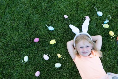 Renkli Paskalya yumurtalarının arasında çimenlerde uzanan tavşan kulaklı mutlu küçük sarışın kız..