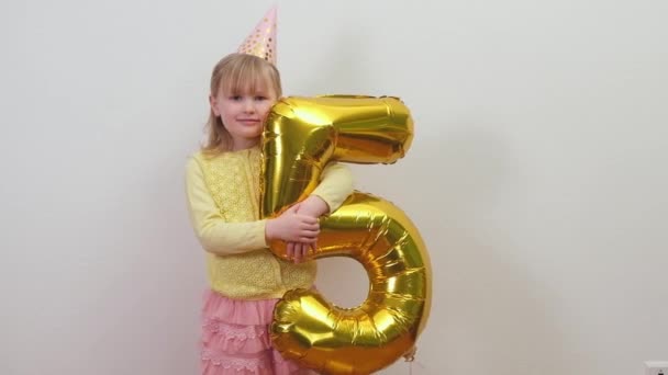 Blondynka biały dzieciak dziewczyna w urodziny kapelusz trzyma złoty foliowy balon 5 i macha do kamery. — Wideo stockowe