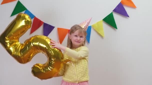 Ευτυχισμένο ξανθό καυκάσιο κορίτσι με καπέλο γενεθλίων κρατά χρυσό αλουμινόχαρτο μπαλόνι νούμερο 5 και χορό — Αρχείο Βίντεο