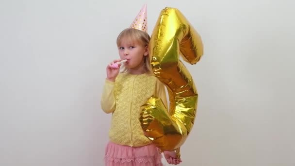 Wenig glücklich Blonde kaukasische Kind Mädchen in Geburtstagshut weht Geburtstagspfeifen und hält Goldfolie Ballon 5. — Stockvideo