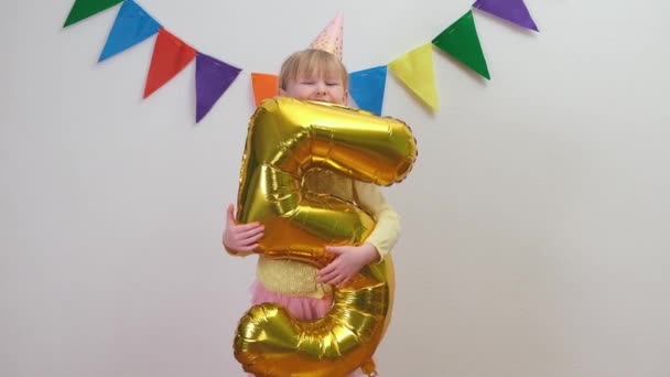 Happy Blonde caucasian kid girl in birthday hat keeps gold foil balloon number 5 and spinning around — Stock Video