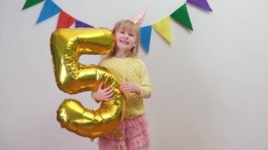 Doğum günü şapkalı mutlu sarışın beyaz kız altın folyo balonunu 5 numarada tutuyor ve eğleniyor..