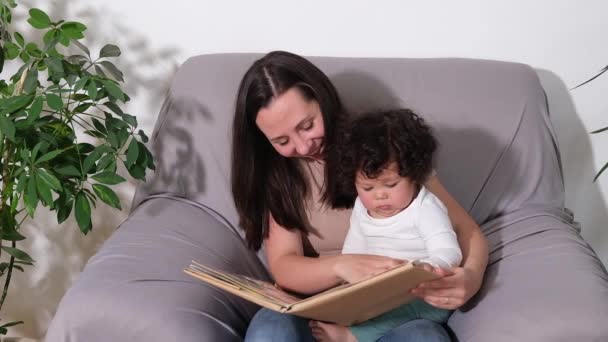 Щаслива мати і змішана раса маленький син читає книгу на дивані вдома . — стокове відео