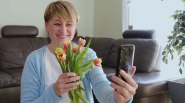 Happy smiling woman holds bunch of flowers and looks at smartphone display and receives messages of congratulation.