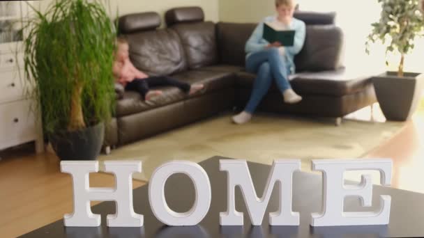 Η μαμά διαβάζει ένα βιβλίο ενώ τα παιδιά διασκεδάζουν στο σαλόνι στο βάθος η επιγραφή HOME. — Αρχείο Βίντεο