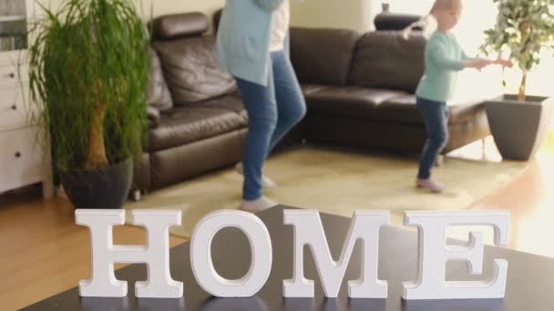 Активна мама і мила маленька дочка танцюють у вітальні на фоні напису Головна — стокове відео