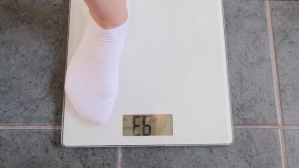 Kvinnliga ben i vita strumpor står på digitala skalor för att kontrollera vikten på golvet i rummet. — Stockvideo