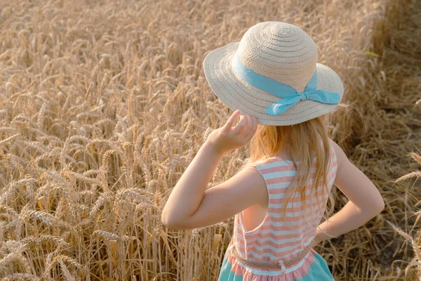 Child blonde girl in straw hat and nice dress holding his hat and smiling at the camera in wheat field