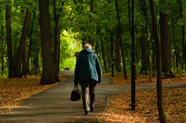 An old hunched woman in pants and a jacket walks along the road in the park. The image of the inevitability of aging clipart