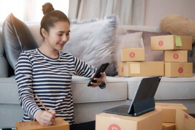 Asyalı küçük işletme sahibi ev telefonuyla siparişleri kontrol ediyor. Ticari perakende pazarı ve çevrimiçi pazarlama dağıtımı, KOBİ e-ticaret kavramı.