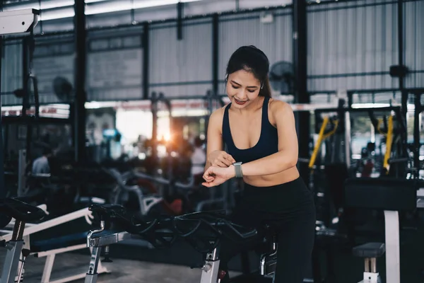 Feliz joven mujer sonriendo y haciendo ejercicio con el deporte en bicicleta de fitness en clase contra la interfaz de fitness. —  Fotos de Stock