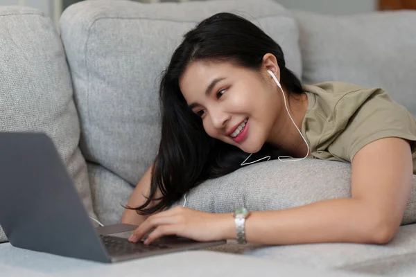 Portrait of Happy Beautiful woman lay down on cozy couch and listen music by streaming on laptop