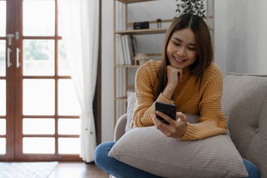 Cheerful asian young woman using mobile phone while sitting on a sofa at home.