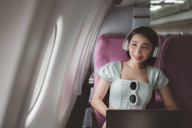 Neşeli Asyalı kadın uçakta dizüstü bilgisayar kullanarak oturur ve seyahat ederken müzik dinler..