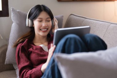 Tablet kullanan ve evdeki kanepede müzik dinleyen çekici, gülümseyen genç bir kadın. yaşam tarzı konsepti.