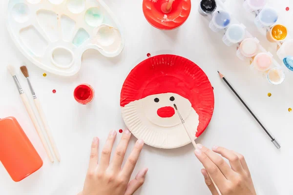 The child glues the parts Santa made from paper plate. Handmade. Project of children\'s creativity, handicrafts, crafts for kids. DIY. Step by step