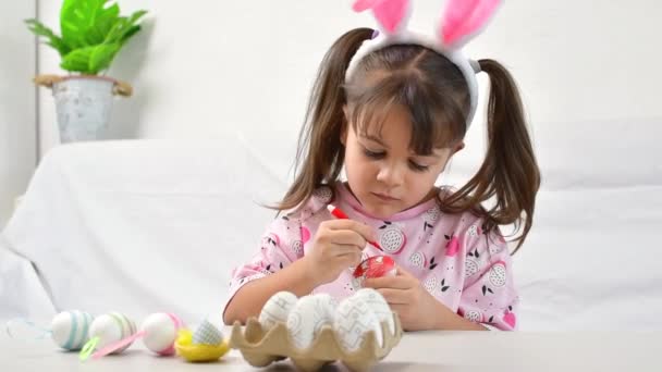 Szczęśliwa dziewczynka z króliczymi uszami maluje jajko włóknem, przygotowując się do szczęśliwego Wielkanocy. — Wideo stockowe