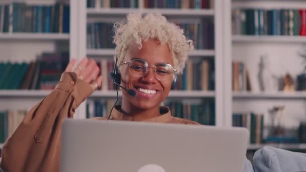 Kulaklık takmış Afro-Amerikalı bir kadın dizüstü bilgisayarın başında kameraya el sallıyor. — Stok video