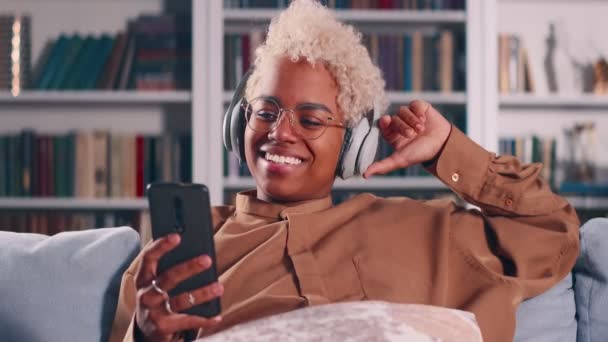Dark skinned woman sit on sofa wear headphones talk to boyfriend by video call — Stock Video