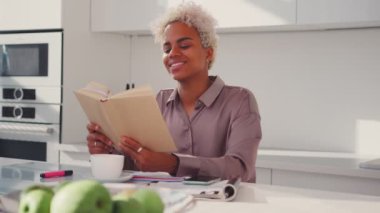 Afro-Amerikalı kadın, elinde açık bir kitapla mutfaktaki masada oturuyor.
