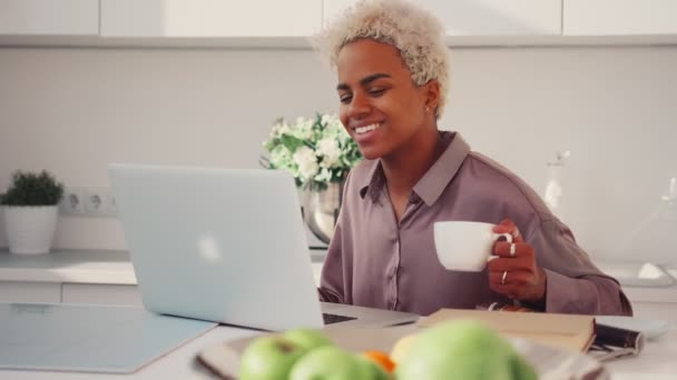 Sonriente mujer afroamericana bebiendo café, mirando la pantalla del ordenador portátil — Vídeo de stock