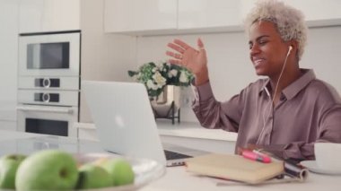 Mutfakta dizüstü bilgisayarla oturmuş gülümseyen Afro-Amerikan kadın.