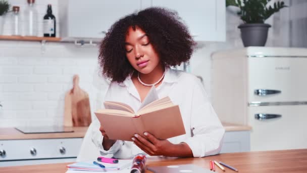 Joven mujer afroamericana leyendo libro mientras está sentada cerca de muebles de cocina — Vídeos de Stock