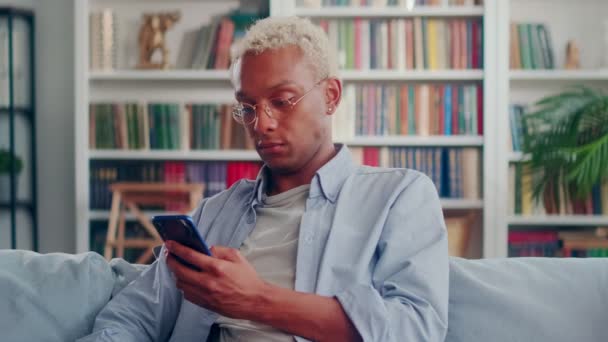 Hombre de piel oscura grave descansando sentado en el sofá y escribiendo en el teléfono inteligente. — Vídeos de Stock