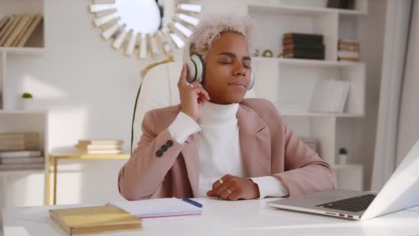 若いアフリカ系アメリカ人の女性がノートパソコンでオフィスでヘッドフォンで音楽を聞く — ストック動画