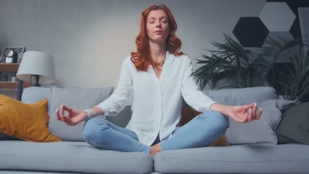 Червоне волосся жінка робить жест мудра, сидячи в положенні лотоса на дивані вдома — стокове відео