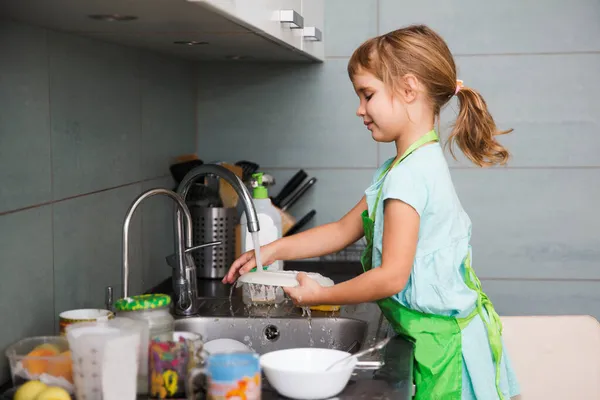 一个可爱的小女孩正在厨房里洗盘子 在现实生活的内部 随意的生活方式摄影系列 帮助父母做家务活的孩子 图库图片